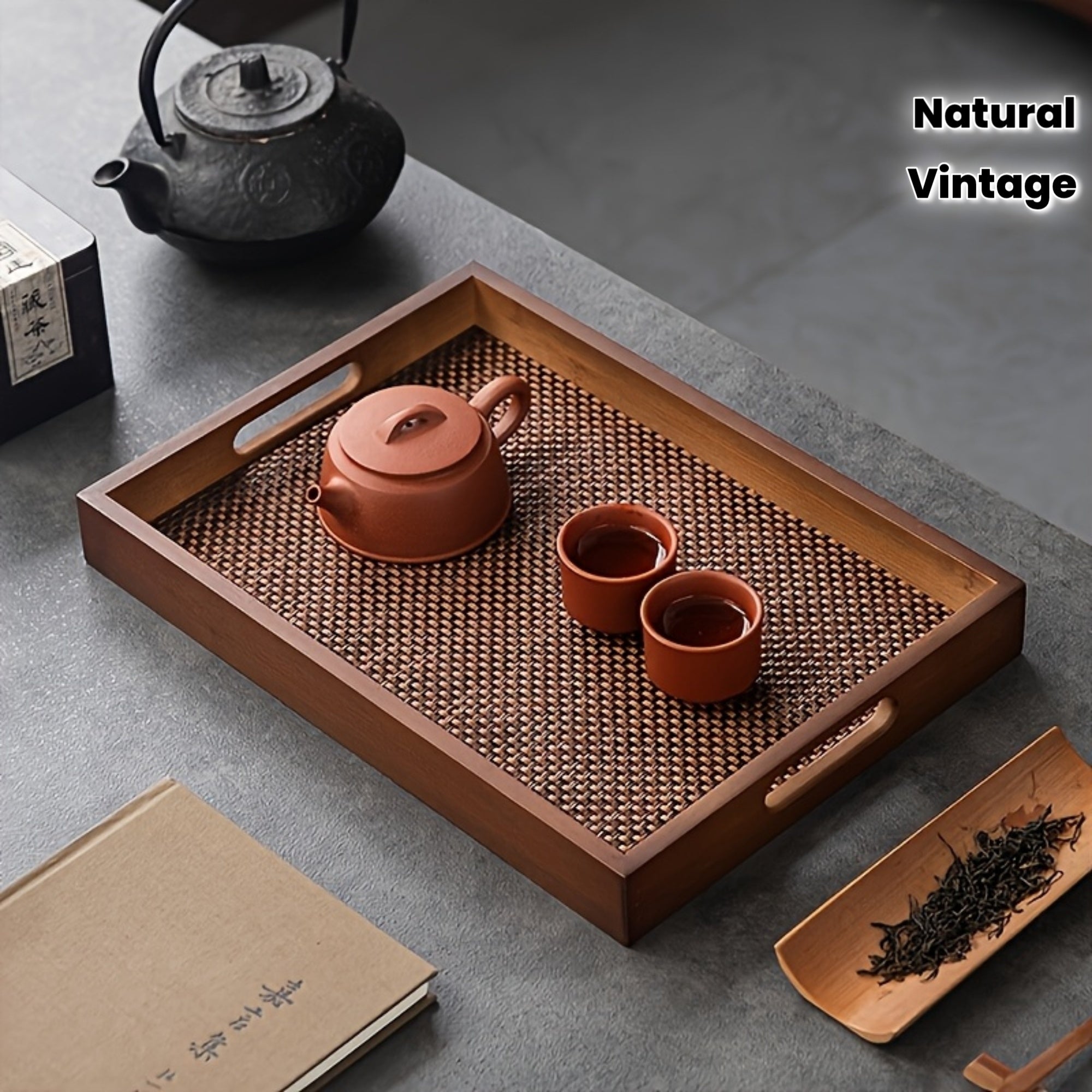 Khây trà gỗ với ấm và chén đất nung màu nâu đỏ, nền bàn xám, bên cạnh là ấm nước đen và sách.