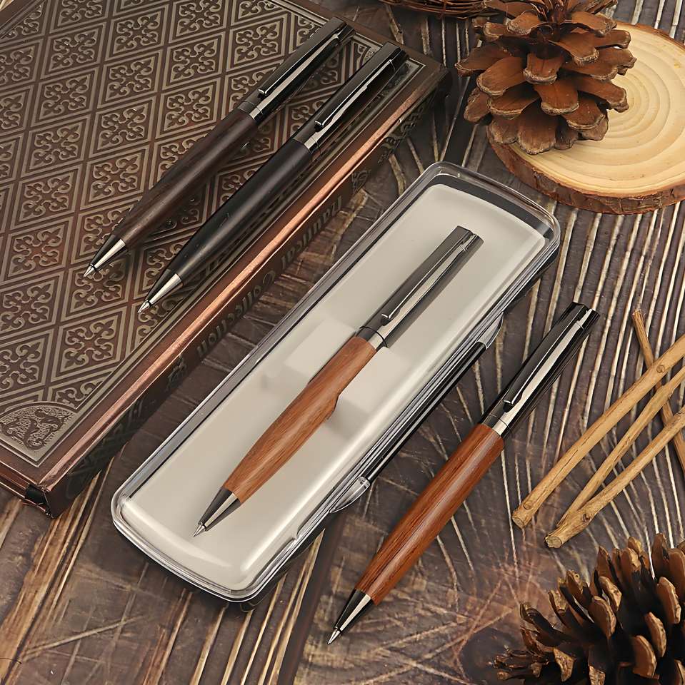 Bộ bút bi gỗ màu nâu và đen trên nền gỗ, kèm hộp đựng trong suốt và quả thông trang trí.