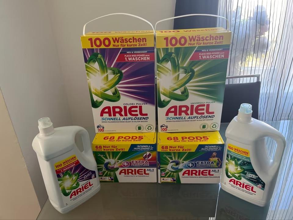 Các hộp và chai bột giặt Ariel màu xanh lá, tím, trắng đặt trên bàn kính.