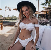 Người phụ nữ mặc bikini trắng, đội mũ rộng vành đen, ngồi trên ghế bên bãi biển nắng.