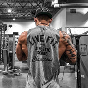 Người tập gym mặc áo xám in "Live Fit," đội mũ, xăm hình, đứng trong phòng tập hiện đại.