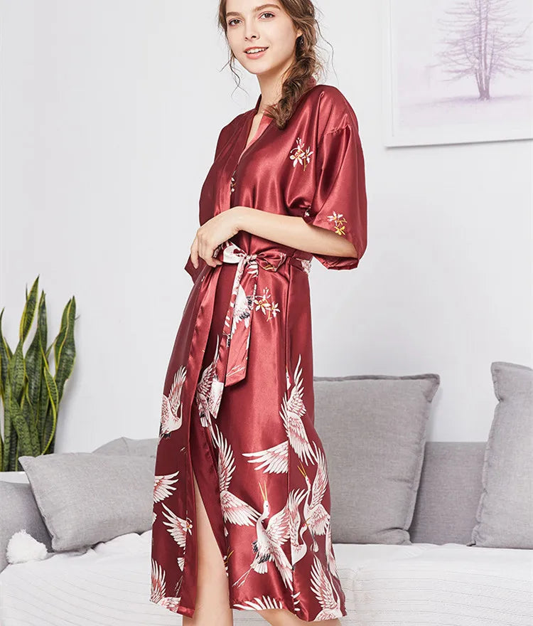 Người phụ nữ mặc váy lụa đỏ in hình chim, đứng cạnh ghế sofa xám, có cây xanh bên trái.