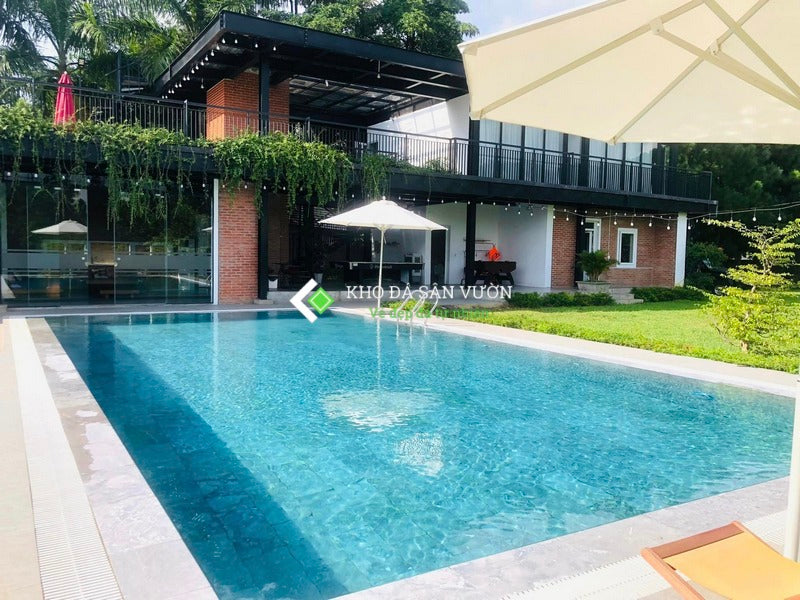 Hồ bơi xanh trong, nhà gạch đỏ, mái hiên đen, cây xanh xung quanh, ô che trắng bên cạnh.