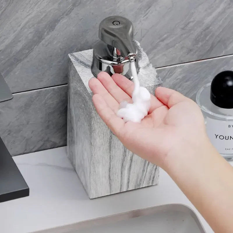 Tay nhận bọt xà phòng từ bình màu xám kim loại, nền gạch xám.