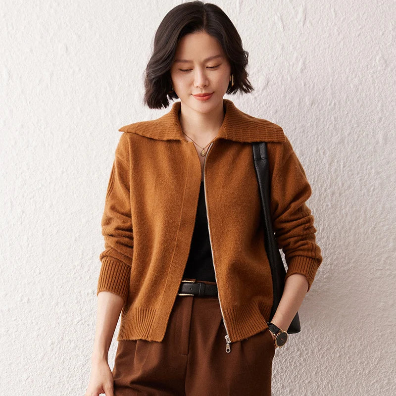 Người phụ nữ mặc áo khoác nâu, tóc ngắn, quần nâu, đeo túi đen, đứng cạnh tường trắng.