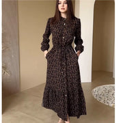 Người phụ nữ mặc váy dài màu nâu đen họa tiết, đứng trong phòng với tường màu be và sỏi trắng.