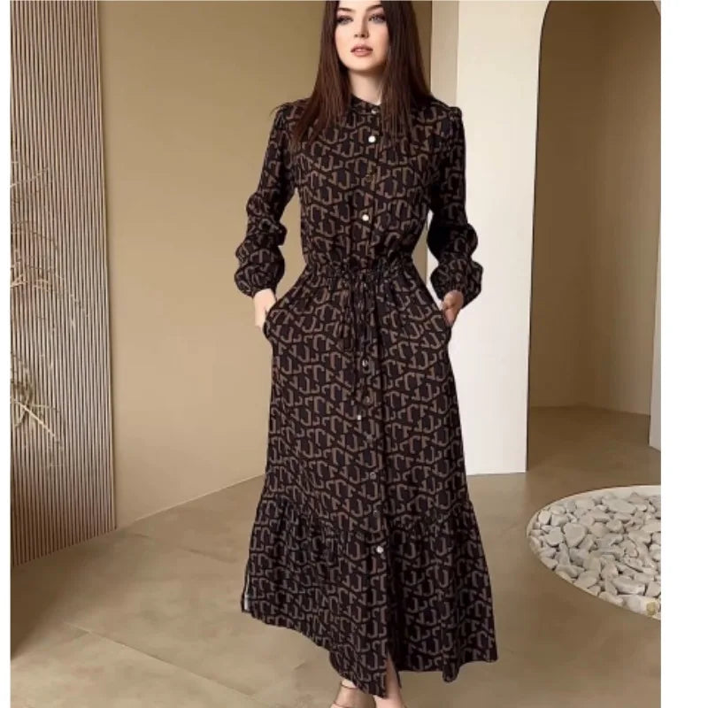 Người phụ nữ mặc váy dài màu nâu đen họa tiết, đứng trong phòng với tường màu be và sỏi trắng.