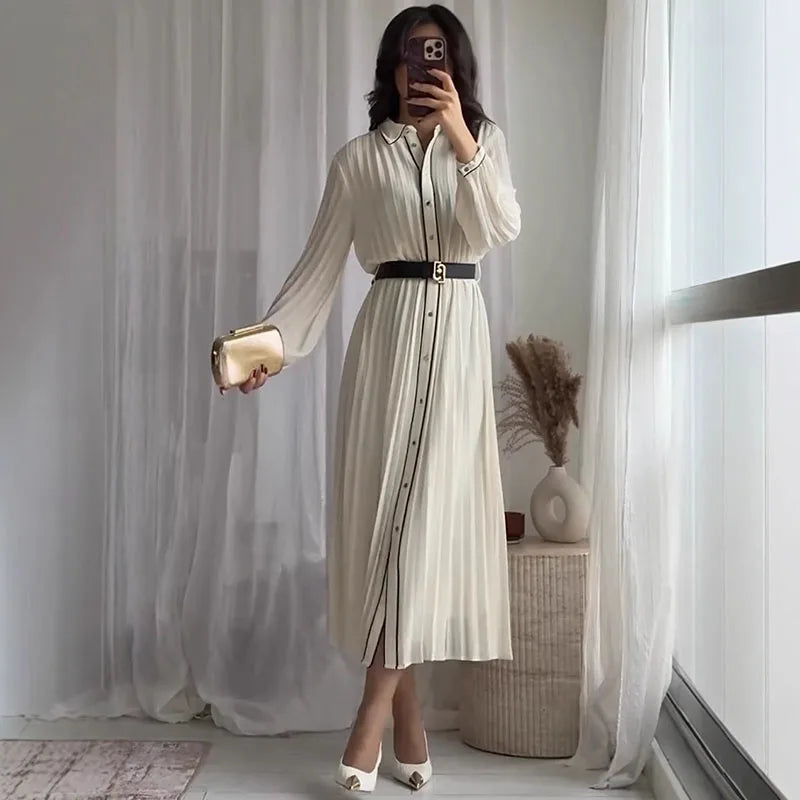 Người mặc váy dài trắng, thắt lưng đen, cầm ví vàng, đứng trước rèm trắng.