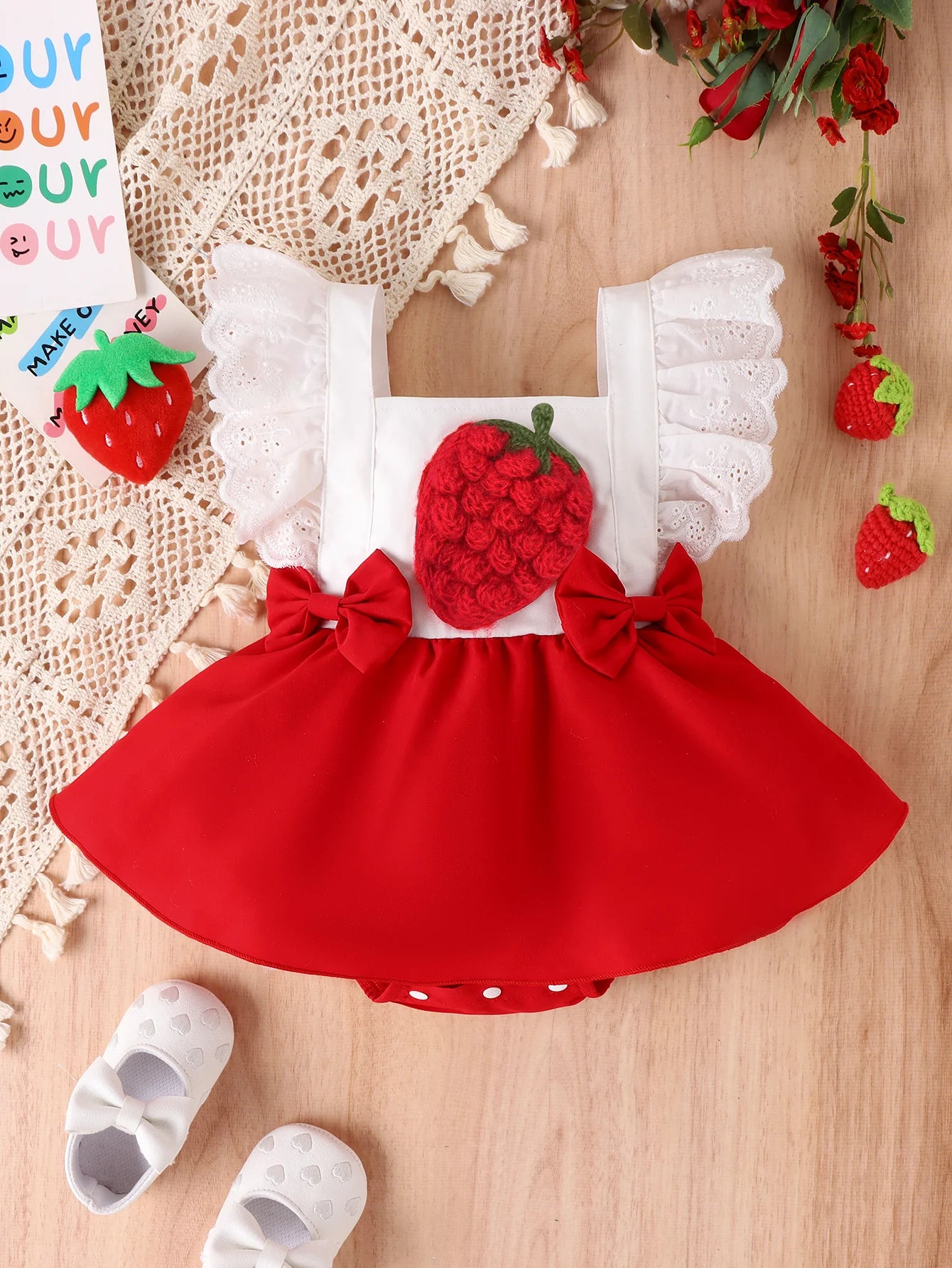 Đầm trẻ em đỏ có hình quả dâu, nơ đỏ, tay áo ren trắng, giày trắng nơ, nền gỗ và hoa đỏ.