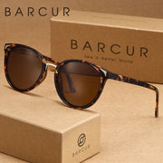 Kính râm nâu gọng đồi mồi, đặt trên hộp giấy màu nâu có logo và chữ "BARCUR" in đậm.