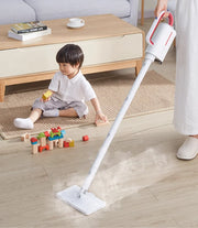 Người dùng máy hút bụi trắng trên sàn gỗ, cạnh bé chơi xếp hình, phòng khách sáng sủa.