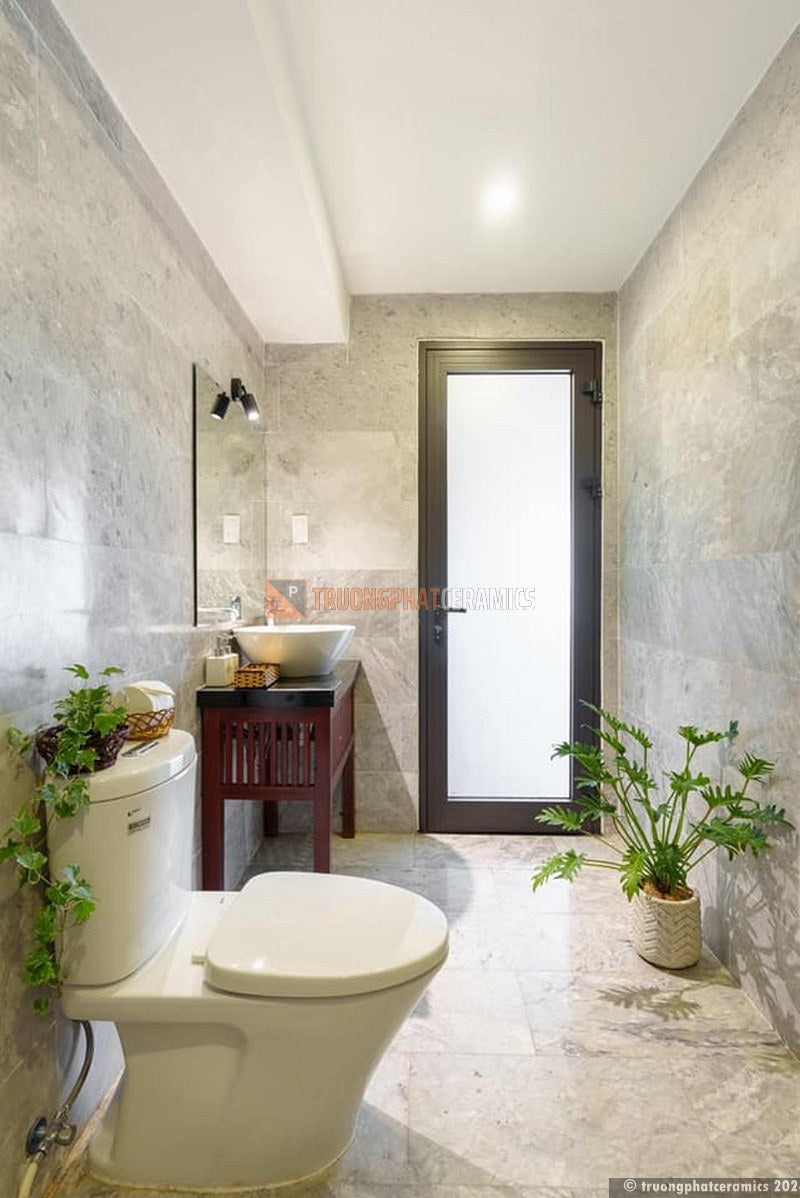 Nhà vệ sinh hiện đại, sàn gạch sáng, tường xám, bồn cầu trắng, chậu rửa, cửa kính mờ, cây xanh.
