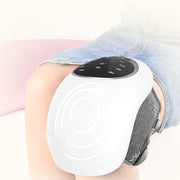 Thiết bị massage trắng trên đầu gối, mặt trên đen có nút bấm, người mặc quần denim xanh nhạt.