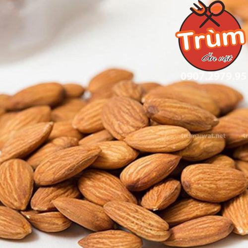 Hạt hạnh nhân màu nâu nhạt xếp chồng lên nhau, nền trắng, có logo tròn màu đỏ ở góc phải trên.