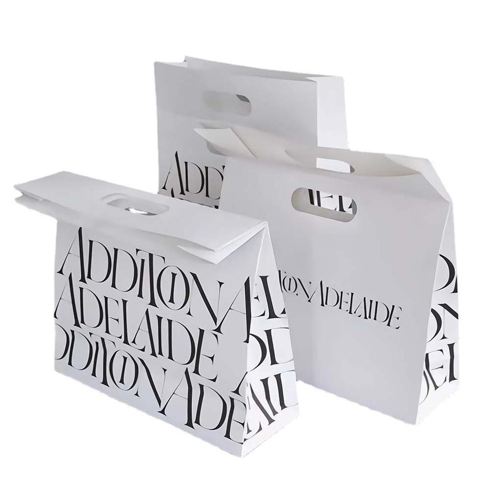 Ba túi giấy trắng có chữ đen "ADDITION ADELAIDE", tay cầm oval, xếp chồng lên nhau.