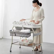 Người phụ nữ chăm sóc em bé trên bàn thay tã màu trắng có họa tiết, bên dưới có giỏ đựng đồ.
