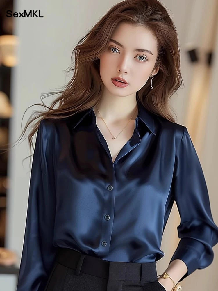 Người phụ nữ mặc áo sơ mi lụa xanh đậm, quần đen, tóc nâu dài, môi đỏ, đeo trang sức tinh tế.