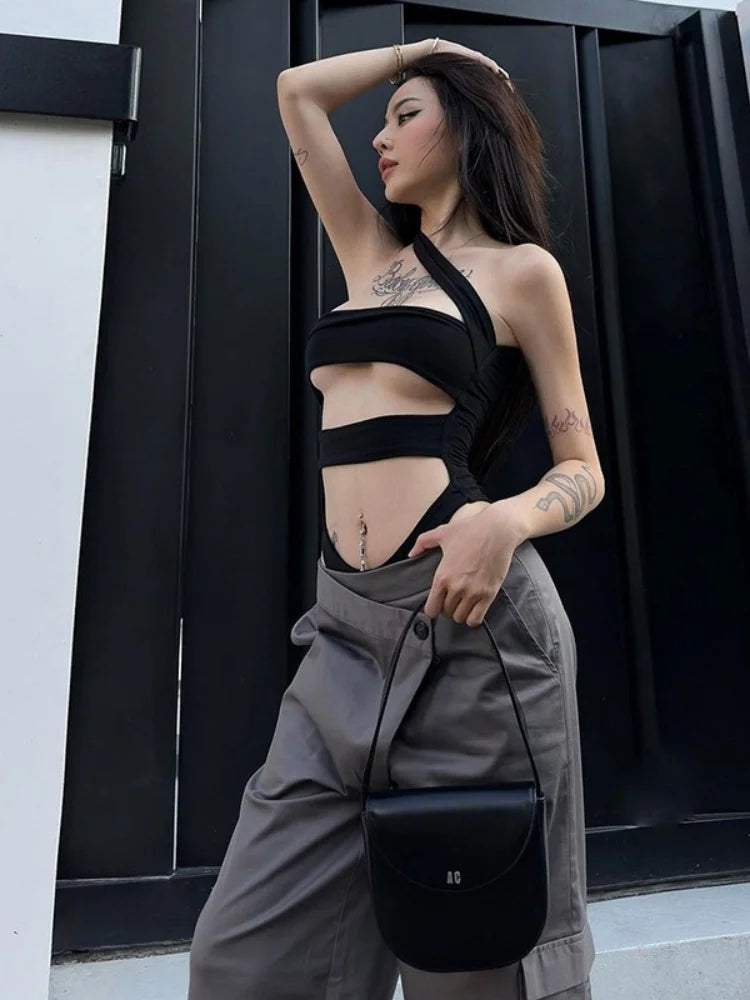 Người phụ nữ mặc áo đen hở eo, quần xám, cầm túi xách đen đứng trước nền đen.
