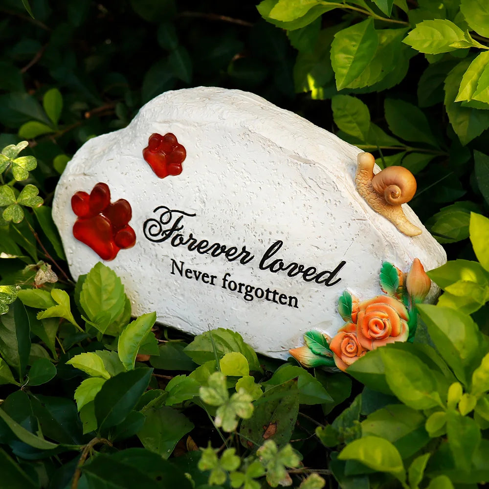 Hòn đá trắng với chữ đen "Forever loved, Never forgotten," xung quanh có lá cây xanh.