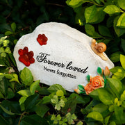 Hòn đá trắng với chữ đen "Forever loved, Never forgotten," xung quanh có lá cây xanh.