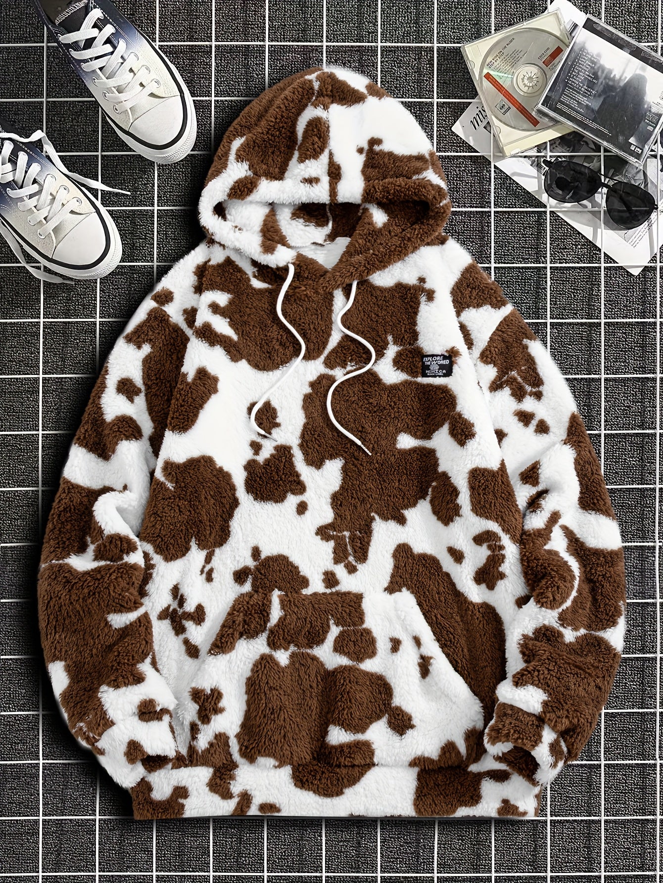 Áo hoodie lông xù trắng nâu họa tiết da bò, đặt trên nền kẻ ô vuông, cạnh giày thể thao và đĩa CD.