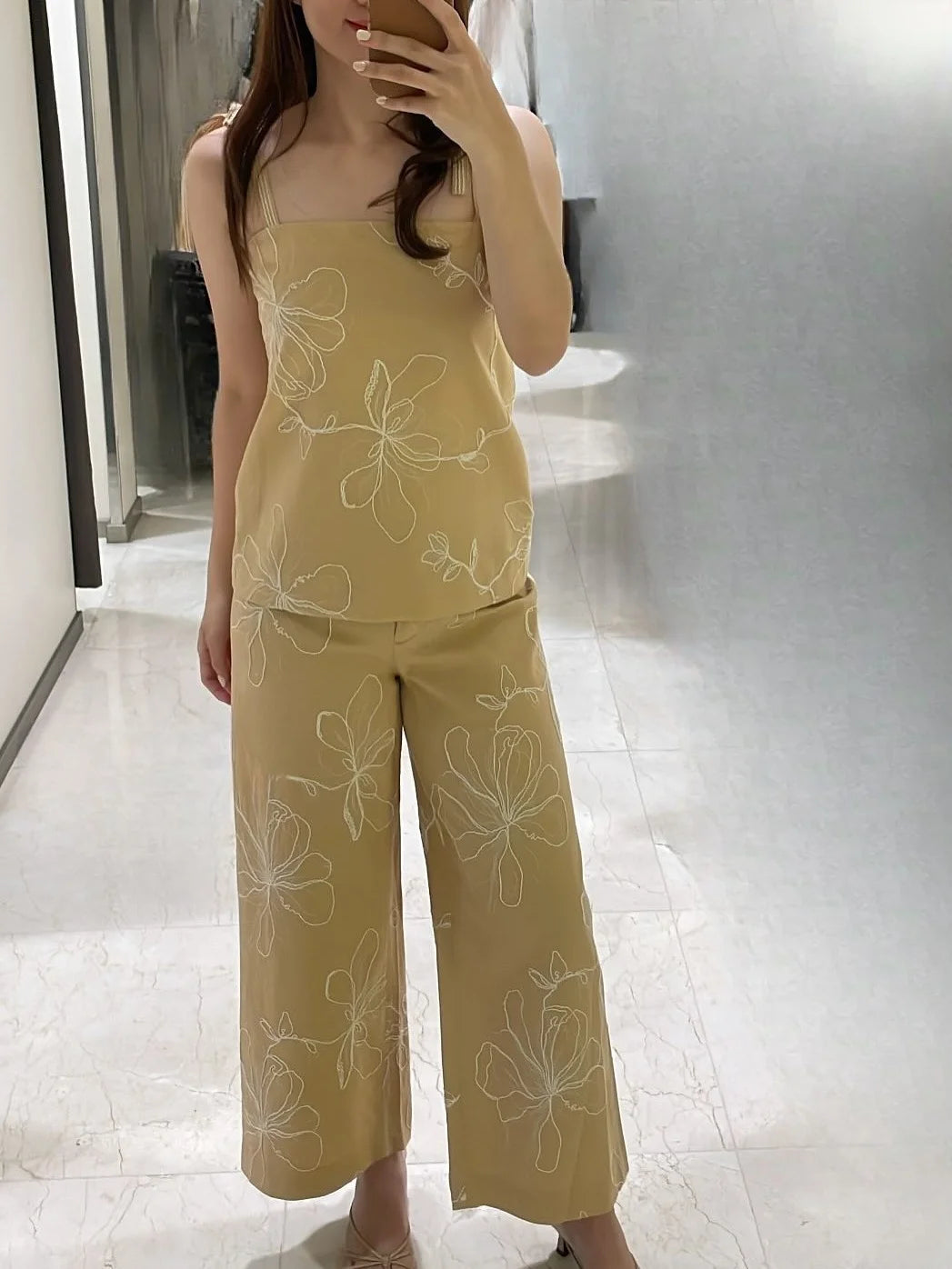 Người mặc bộ đồ màu be với hoa văn trắng, áo hai dây và quần dài ống rộng, đứng trên nền gạch sáng.