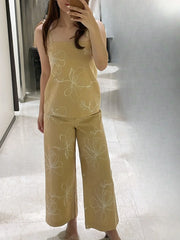 Người mặc bộ đồ màu be với hoa văn trắng, áo hai dây và quần dài ống rộng, đứng trên nền gạch sáng.