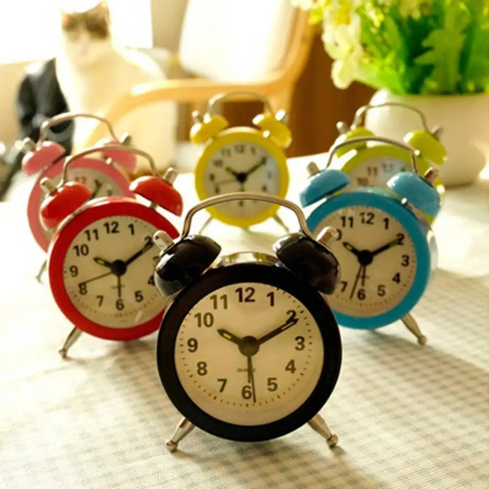 Sáu đồng hồ báo thức nhiều màu trên bàn kẻ caro, nền mờ có hoa và mèo.