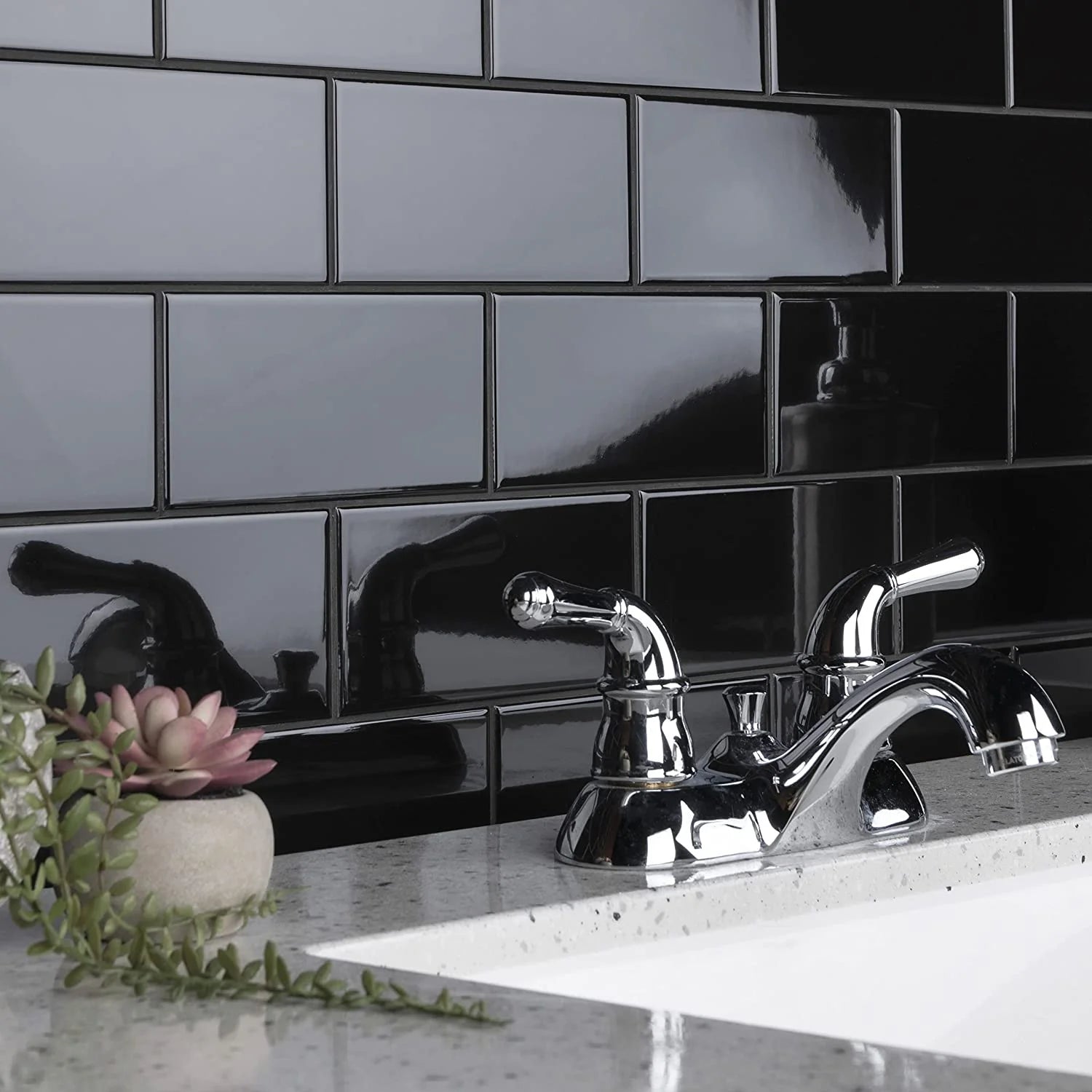 Vòi nước kim loại sáng bóng, gạch ốp tường màu đen, chậu cây nhỏ trên bề mặt đá trắng.