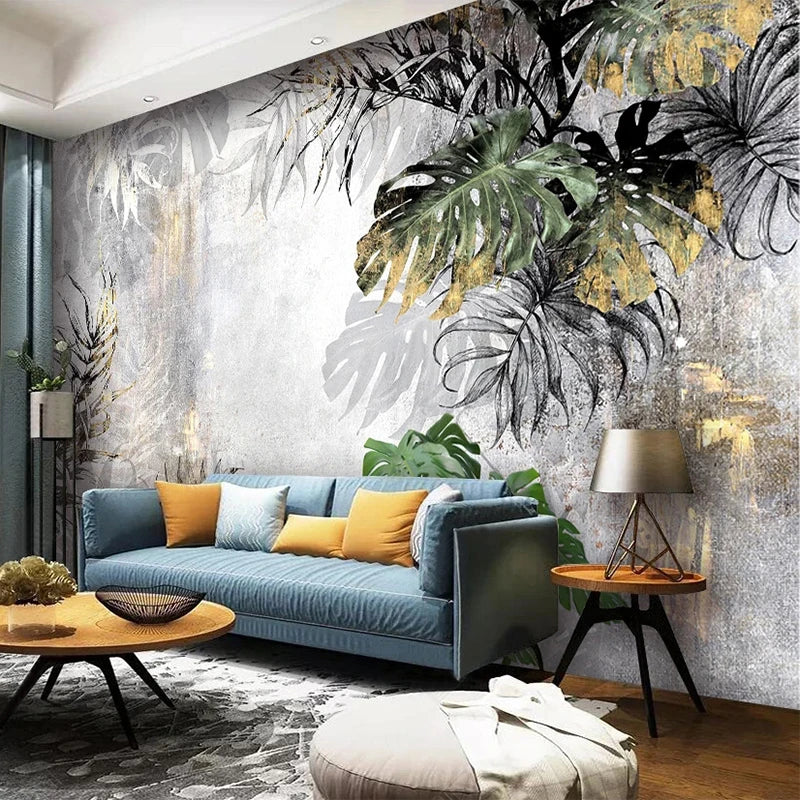 Sofa xanh với gối vàng, nền tường họa tiết lá lớn, bàn gỗ tròn, đèn bàn, sàn gỗ.