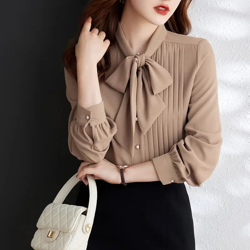 Người phụ nữ mặc áo sơ mi nâu có nơ lớn, cầm túi xách trắng, nền tối và tường trắng.